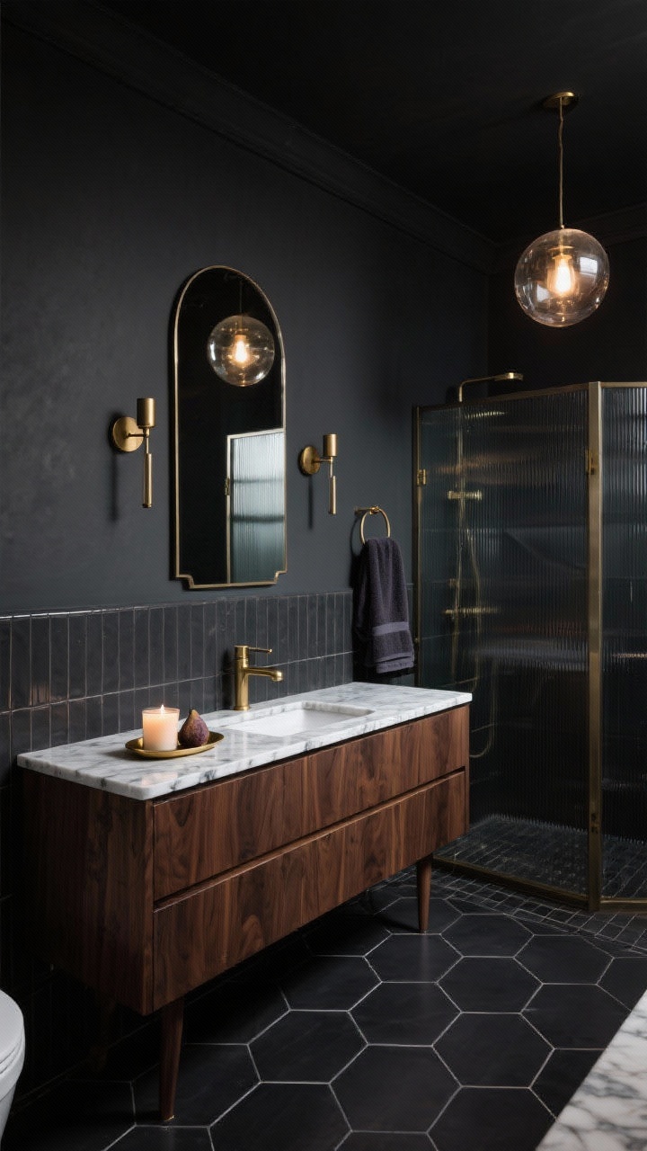Wide, moody scene of a dramatic bathroom with deep charcoal walls and glossy graphite tiles: a sleek walnut vanity topped with a marble-look counter featuring white veining, a large arched mirror, brushed brass wall sconces and faucet, black hex floor tiles, a smoked glass shower screen, and a statement globe pendant casting a low, atmospheric glow; plush dark towels and a fig-scented candle on a small brass tray for finishing detail.